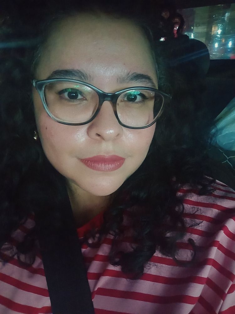 mulher branca de cabelos castanhos e cacheados, usando uma camiseta rosa com listras vermelhas e óculos de grau. tô dentro do carro, então aparece o cinto 