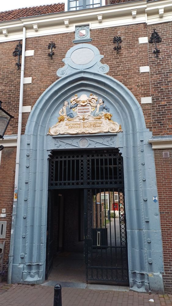 Blauwe poort in een bakstenen muur. Boven de doorgang een reliëf met Latijnse tekst. In de poort een ijzeren hekwerk met deur. Daarachter een donkere doorgang naar een binnenplaats.