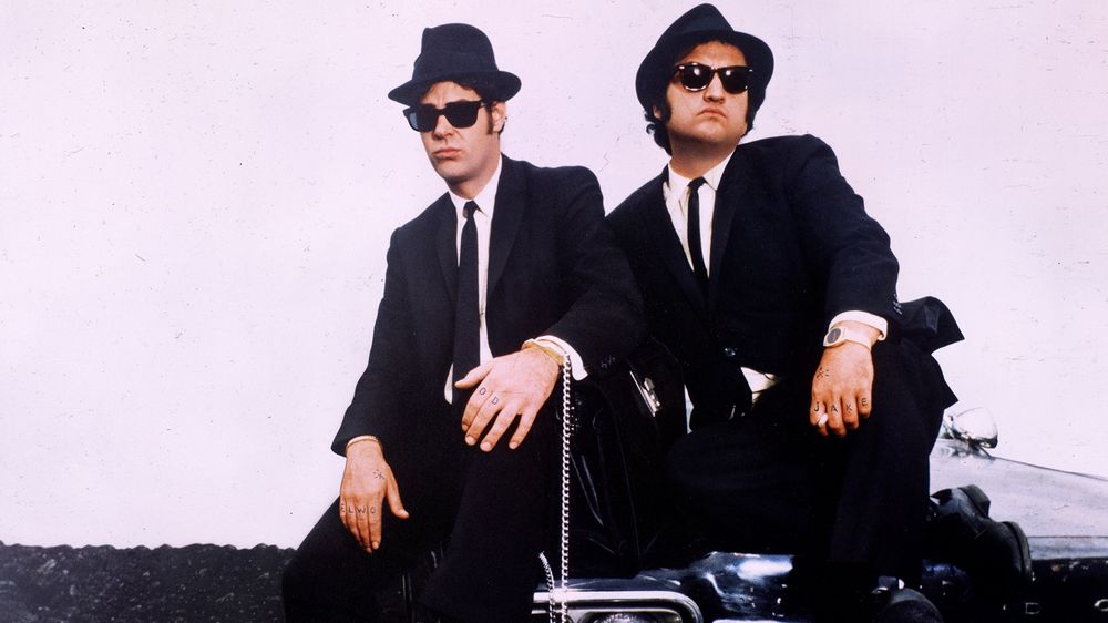 Two men in suits, fedoras, and sunglasses sitting on the hood of a car that's got a cop motor, a 440-cubic-inch plant. It's got cop tires, cop suspension, cop shocks. It's a model made before the catalytic converter so it'll run good on regular gas.