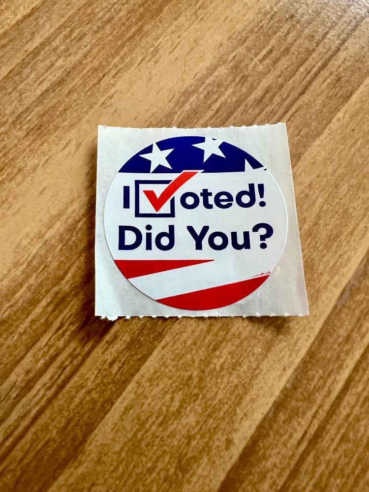 A circular sticker reading “I voted! Did you?” On a wax backing sitting on a wooden table.