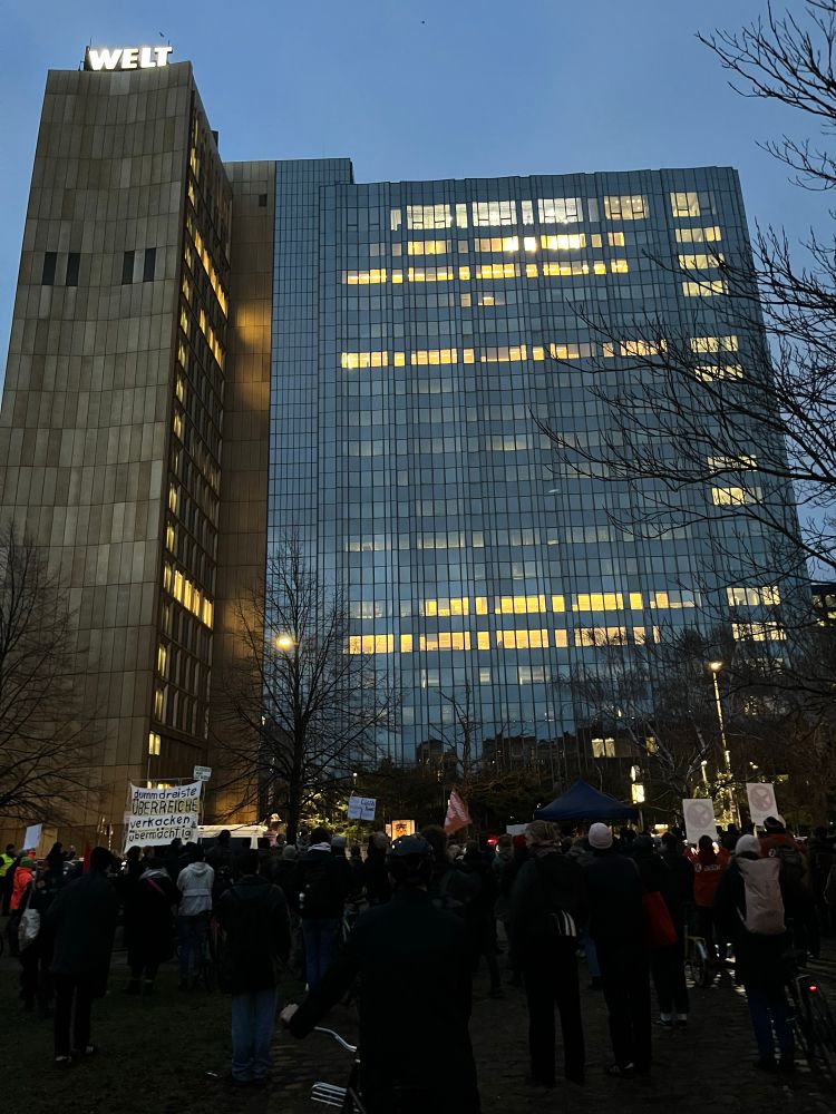 Hochhaus des Springer Gebäudes. Im Vordergrund Demonstrierende mit Transpis.
