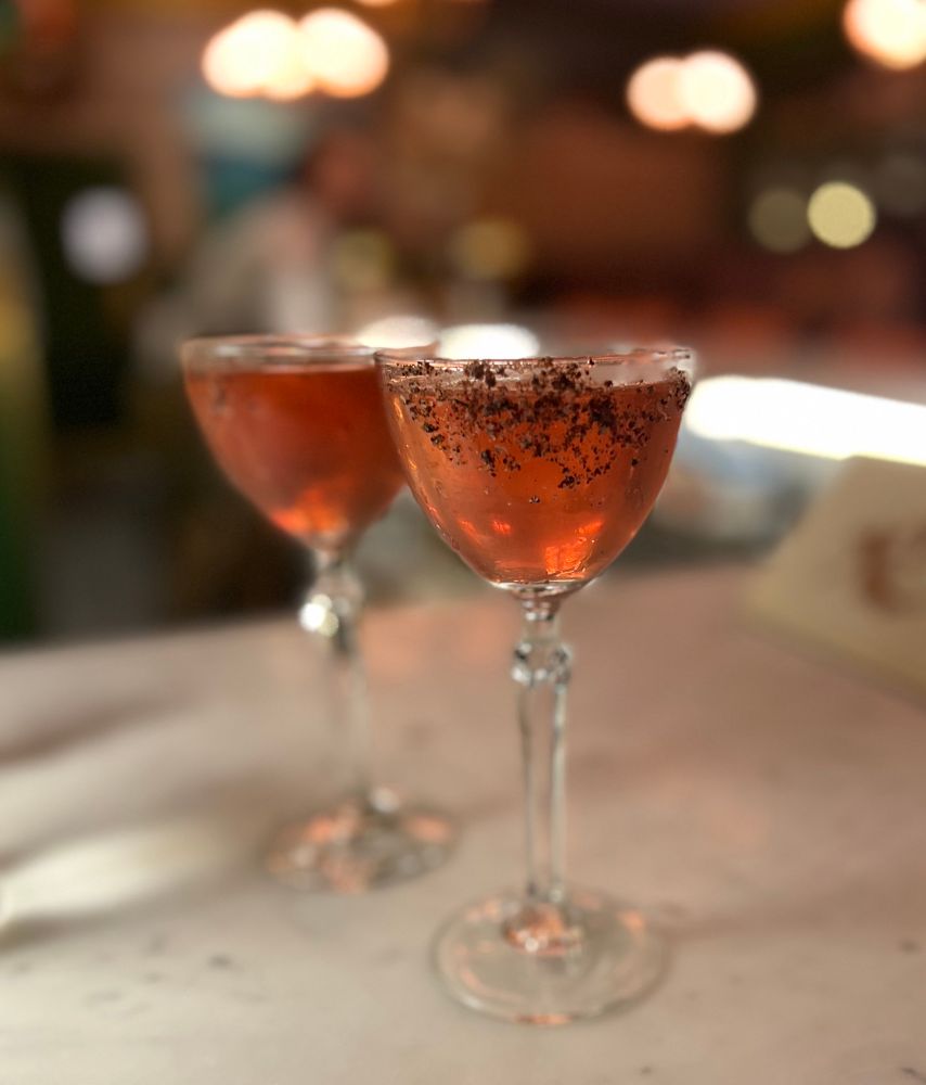 Two glasses of a pinkish-purple martini made with Kalamata olive brine. The glasses sit atop a marble bar.