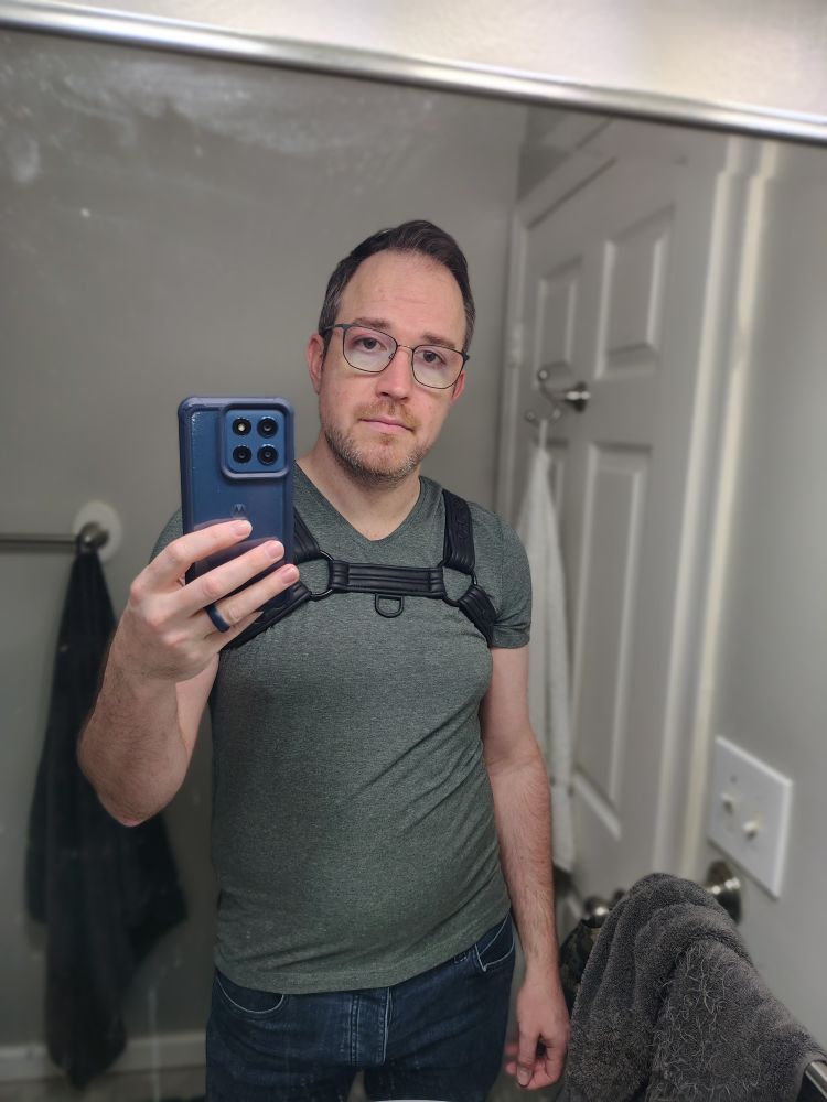 Joe, a white man with brown hair and stubble, wearing jeans and a green shirt with a neoprene harness over it