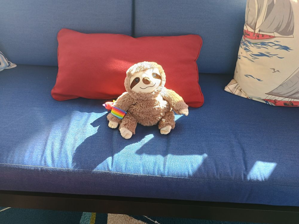 A plush sloth wearing a rainbow wristband, sitting alone on a blue sofa