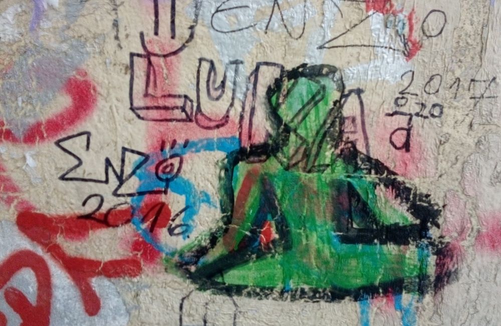 Bild zeigt Graffiti mit sitzender umrisshaft dargestellter grüner Figur, schwarz umrandet und umgeben und zum Teil überschrieben von anderen Graffitis, Worten und Tags.