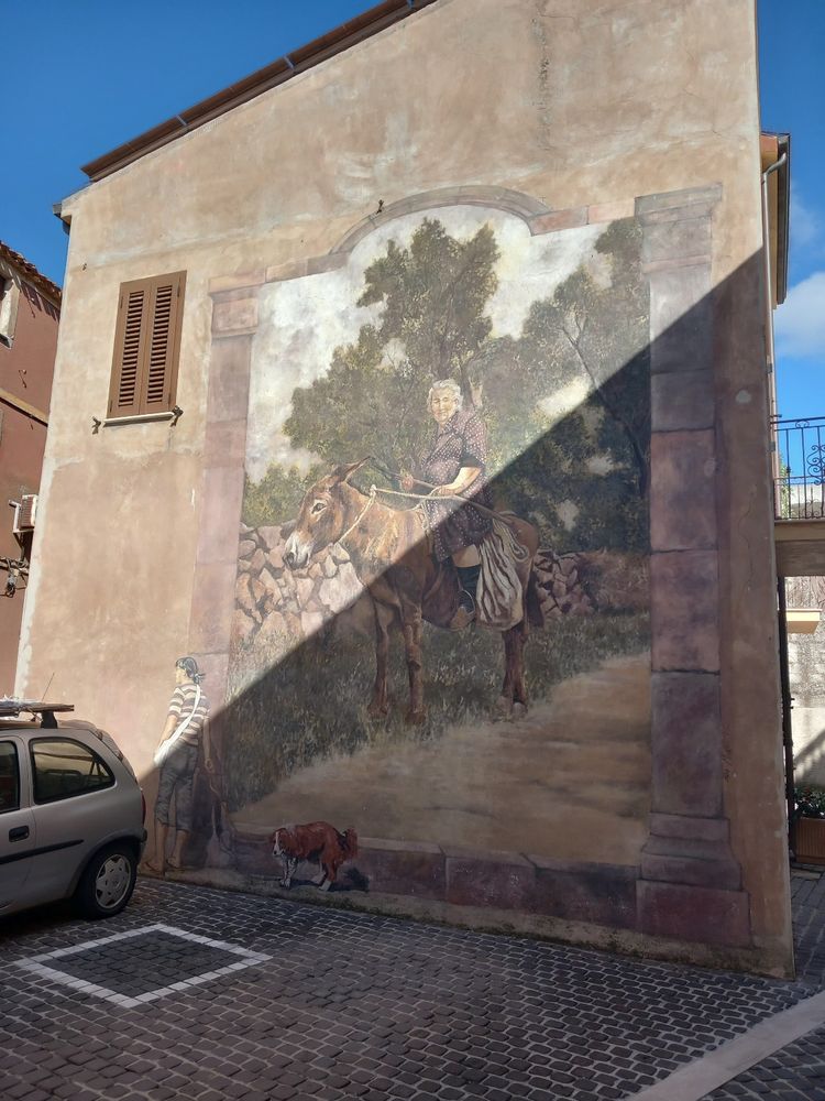 Murales of a woman walking her dog in front of a portrait of a Sardinian woman riding a donkey on a country pathway.
