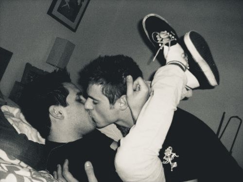 Two men kissing. One is on his back with both ankles crossed behind his partner’s head and his hand loosely clasped behind his ear. His partner is leaning over and holding him 