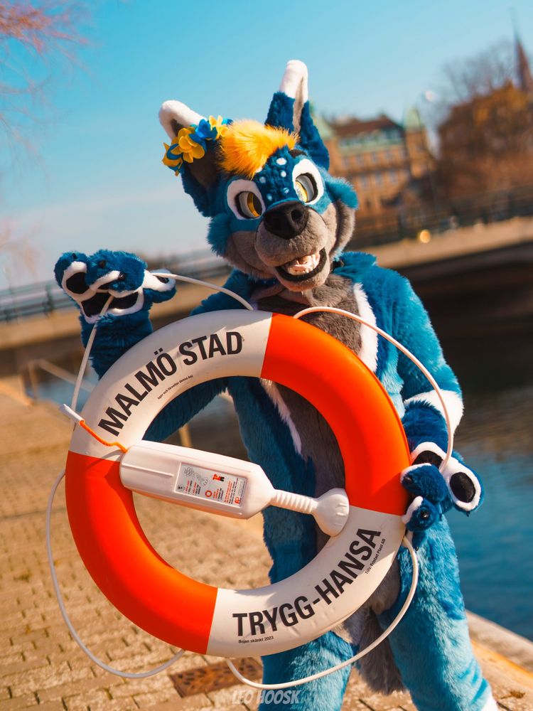 A blue and gray fursuiter named Vextrum holds a lifebuoy near the water, smiling playfully.
