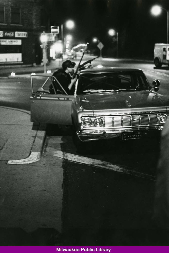 Black and white photo of two police officers with Huns drawn crouching alongside a squad car on a city street