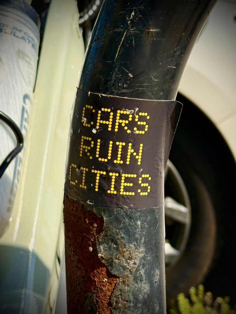 Sticker that reads “cars ruin cities” applied to a rusting metal bike rack with a water bottle and a car visible in the background