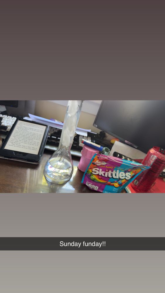 A picture that shows a kindle, a bong, skittles - wild berry and tropical mix, a pink grinder, a blue lighter and a red monster on a brown desk with some computer monitors and a keyboard. The picture says “Sunday Funday!!” Like the caption 