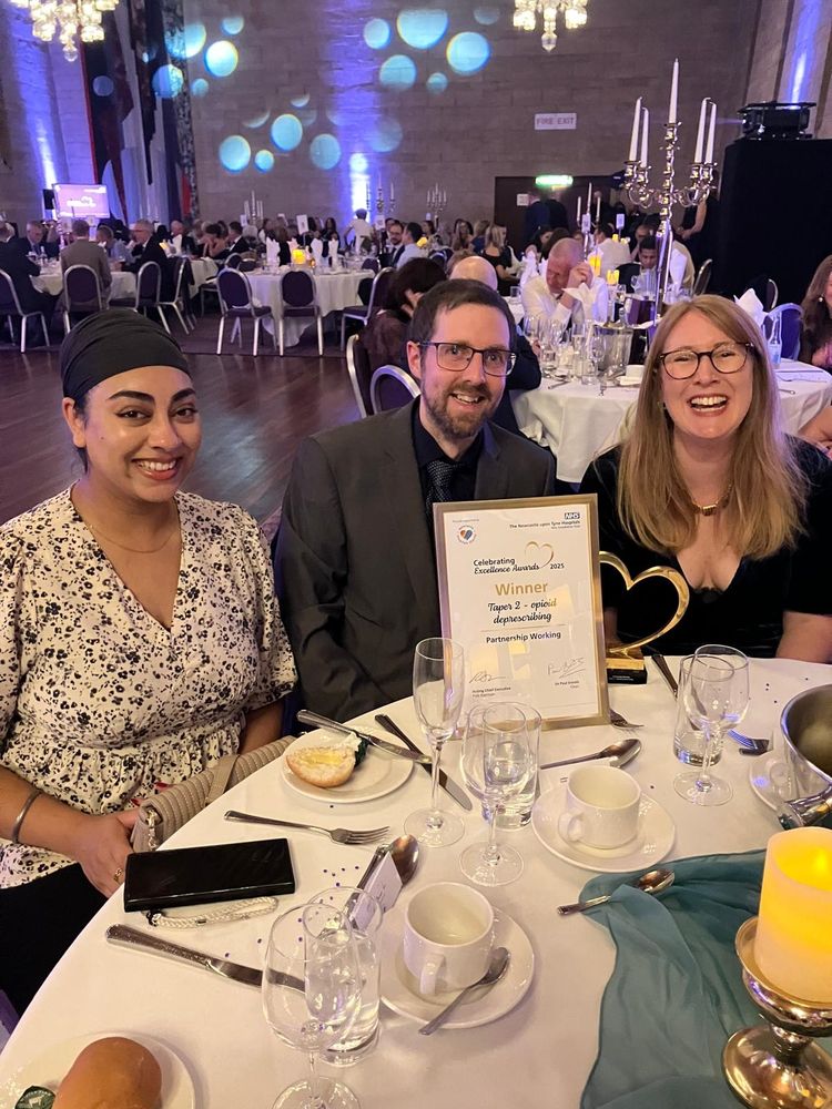 Kiran, Thomas, Ang sat at a dinner table, with the awards ceremony in the background. On the table is the award for TAPER2. 