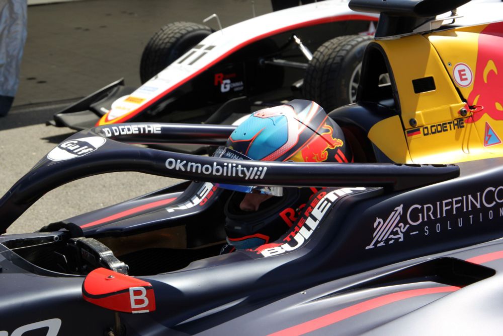 Close-up of Oliver Goethe, wearing his Red Bull liveried crash helmet, in the cockpit of the dark blue and yellow Red Bull liveried MP Motorsport F3 car in the support race paddock at Imola. The number 11 Campos car can be seen in the background.