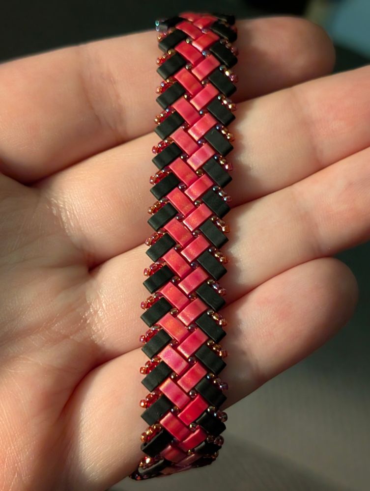 A herringbone half-tila beaded bracelet in matte black and red. It is accented with iridescent transparent red seed beads.