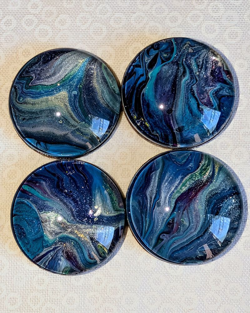 A set of four circular, one-inch magnets. They're primarily shades of blue with swirls of silver, green, yellow and shades of purple. They were made using glass cabochons that have been dipped in wet fluid art (a type of abstract painting).
