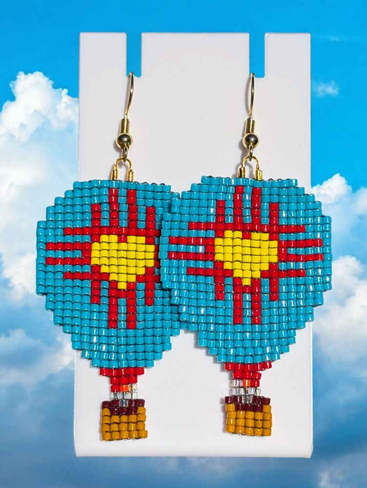 A pair of beaded Zia heart hot air balloon earrings over a cloudy, blue sky background. The earrings are mainly turquoise with the Zia shape in red and a yellow heart center.