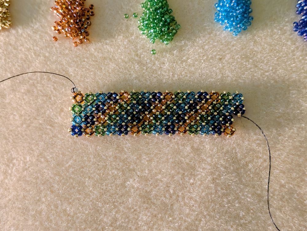 A partially finished beaded bracelet in orange, green, blue, purple, and gold seed beads. Above it are little piles of each color of beads that I'm working with.