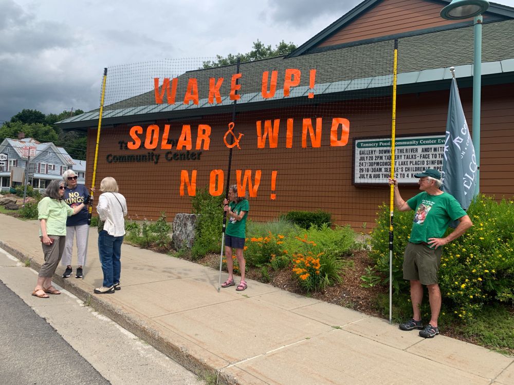 Three people stand on the sidewalk outside the Tannery Pond Community Center displaying a large banner that reads Wake Up! Solar & Wind Now!