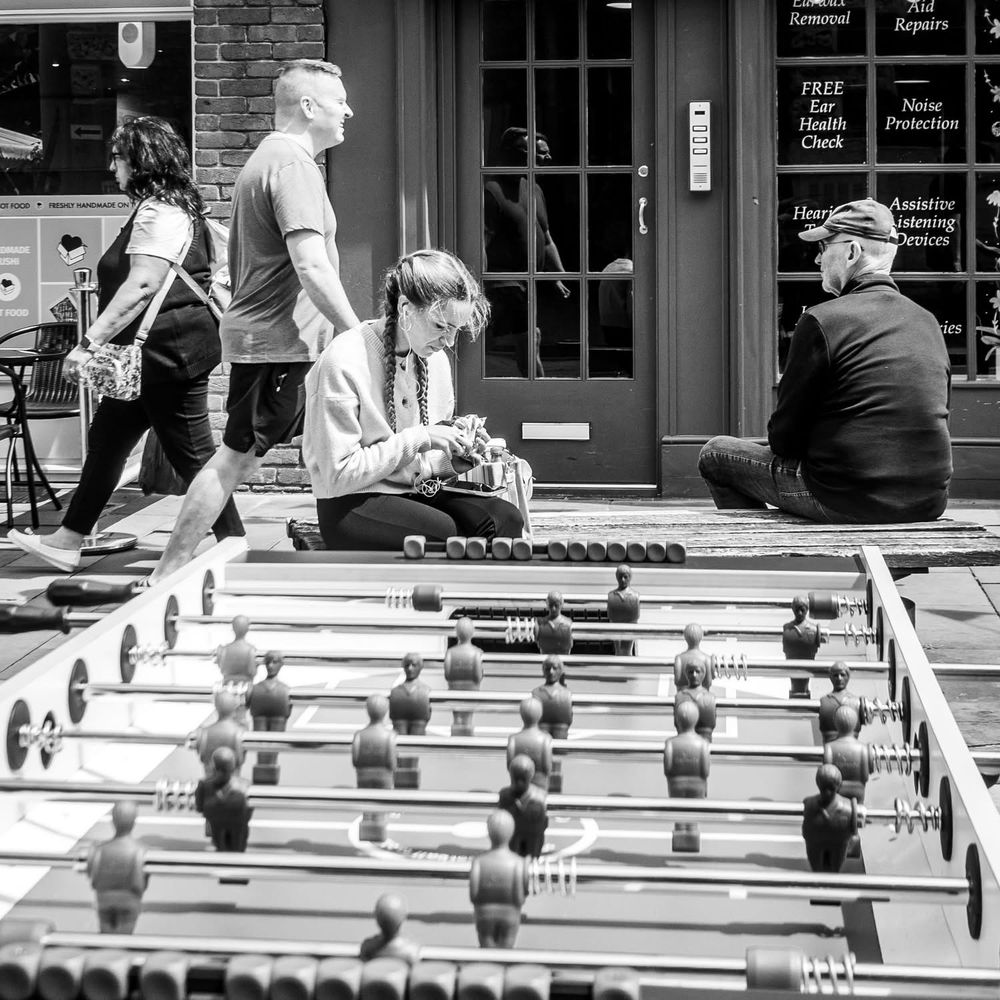 A street photograph of a view over a street table football game of a girl sat on a street bench. There is also a man in a cap with his back to me sat on the bench. Black and white photo. 