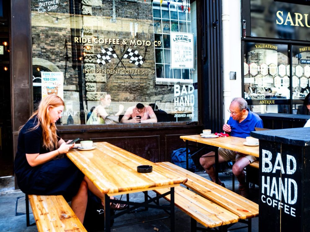 Outside seating of a coffee shop two people sit on separate tables looking into their phones. Inside the coffee shop two people can be seen through the window also looking into their phones. Colour photograph 