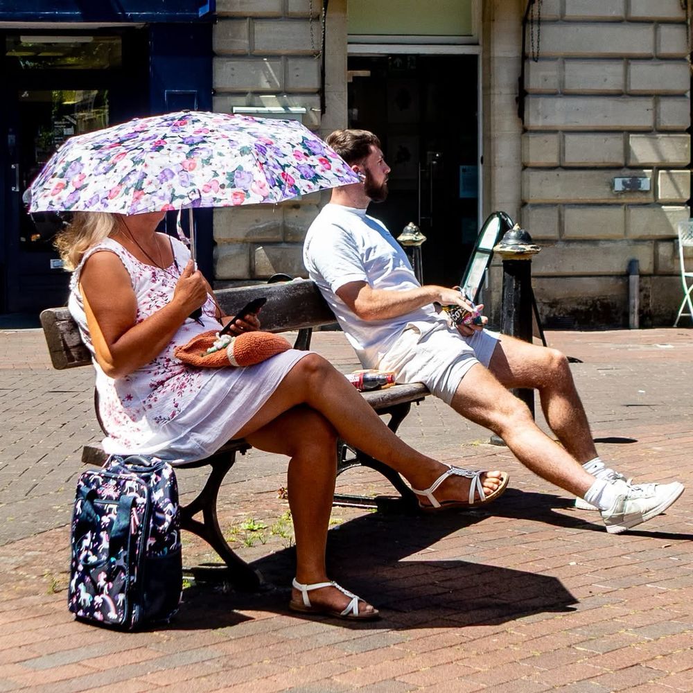 A lady sat on a street bench in a summer dress holding a parasol low down so that it hides her face. There is a man in shorts sat at the other end of the bench. Colour photograph. 