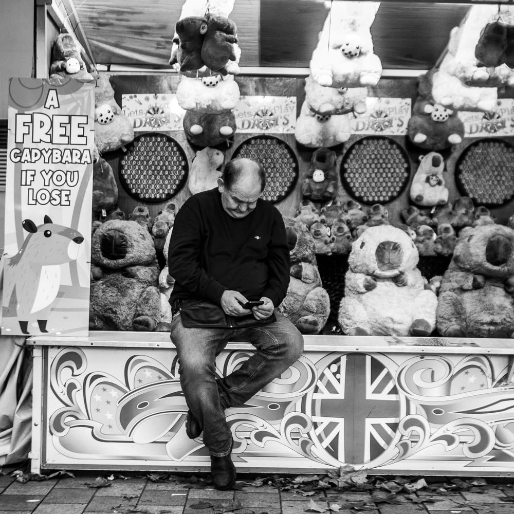 Fair stall attendant sat on the front of his attraction looking down at his phone. Black and white photo. 