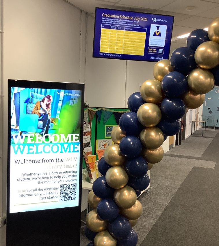 A digital Welcome screen is shown beside part of the balloon arch, made of gold and blue balloons