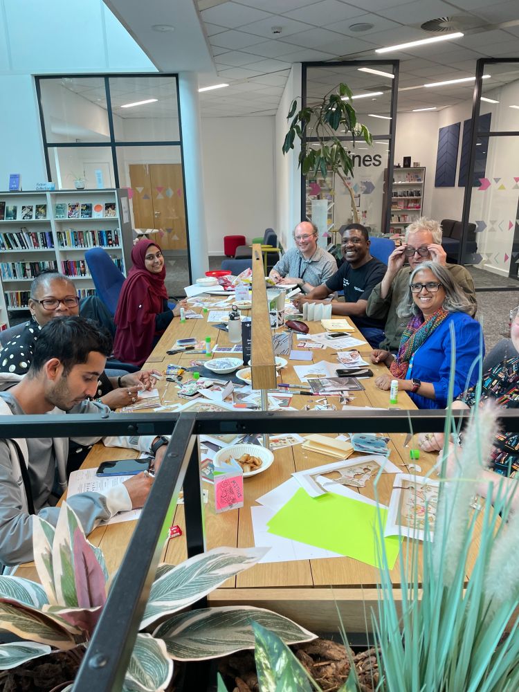 A photo of 8 people sat around a table with craft supplies on making zines.