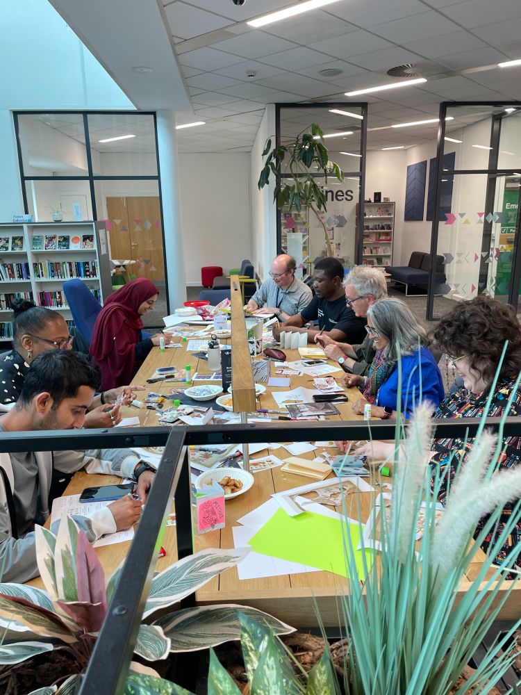 A photo of 8 people sat around a table with craft supplies on making zines.