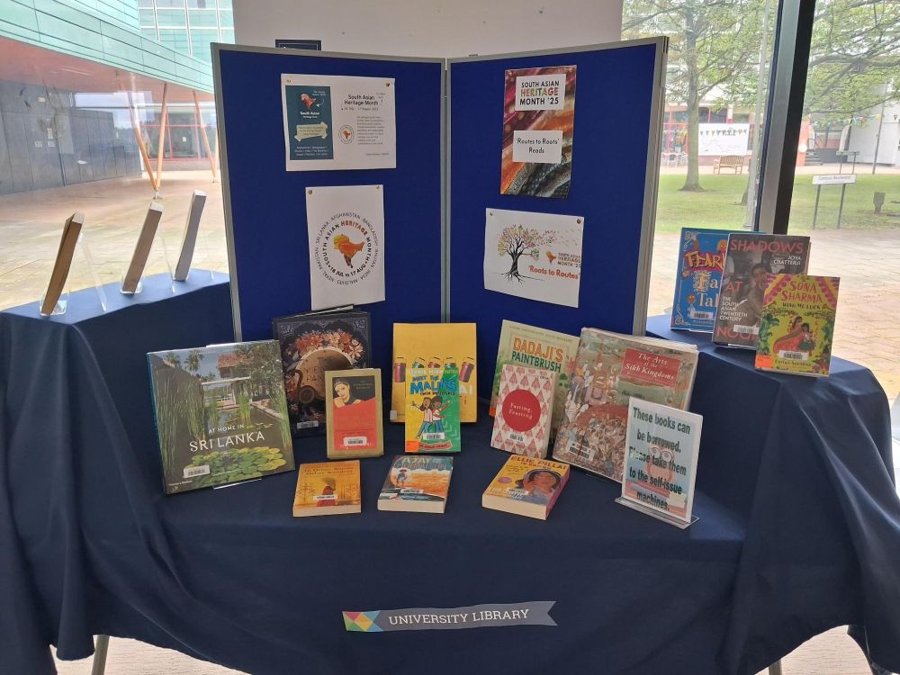 A photo of a table with books on display about south asia and south asian heritage, with a board in the background with posters about South Asian Heritage Month: Routes to Roots.