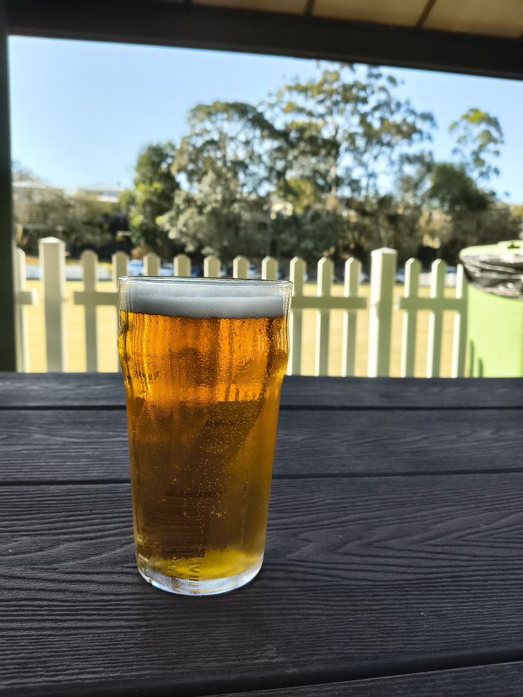 Pint of cold lager on a table with a bowling green and trees behind a white picket fence.