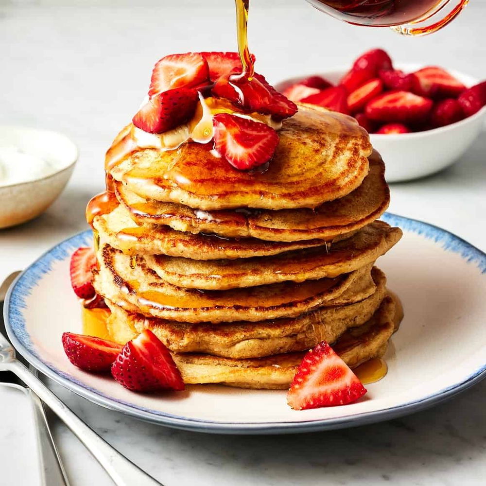 A stack of seven oatmeal pancakes with sliced strawberries on a white plate with a blue lip. Golden maple syrup is drizzling over the pancakes. In the background there is a bowl of sliced strawberries.