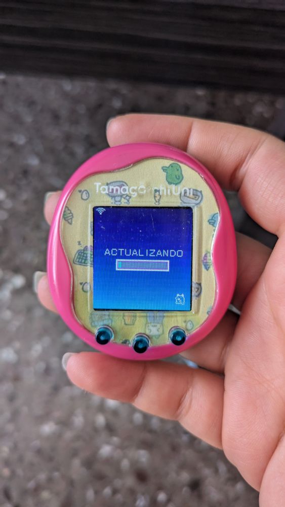 Tamagotchi uni rosa con el faceplate (el papelito que va detrás del plástico que va por encima de la pantalla) personalizado, amarillo con dibujitos de personajes de tamagotchi. En la pantalla se ve "ACTUALIZANDO", la barra de progreso y el sprite de Mametchi corriendo para que sepas si sigue vivo o se ha colgado, supongo.