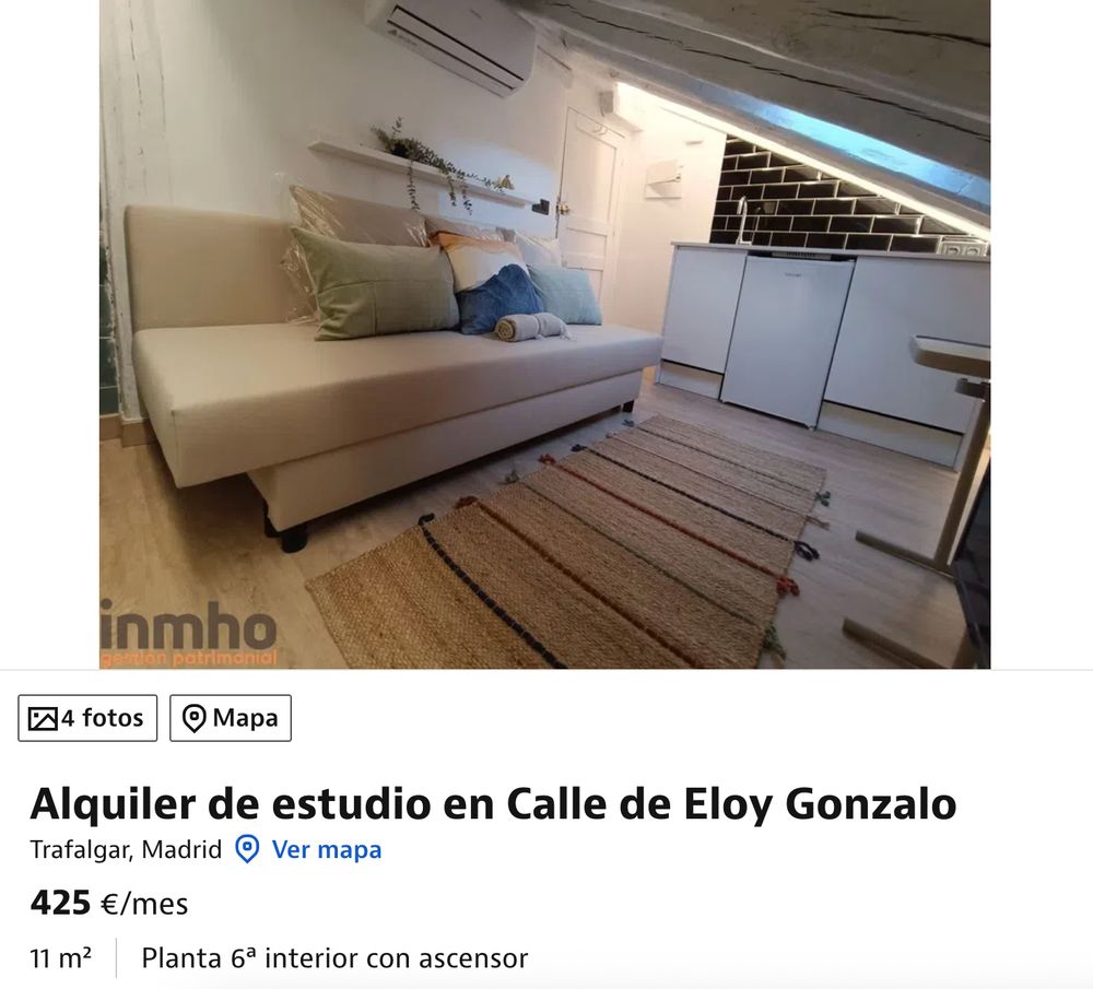 Foto de una estancia de un piso, se ve un sofá en primer plano, con una alfombra delante. Al fondo se ve el grifo de la cocina y el lavavajillas. Los techos son abuhardillados. Debajo se lee "Alquiler de estudio en Calle de Eloy Gonzalo, Trafalgar, Madrid. 425€/mes. 11m², Planta 6ª interior con ascensor"