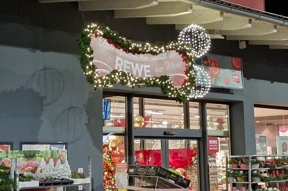 Rewe Weihnachtsdekoration. Ein Schild auf dem steht "Frohe Weihnachten wünscht dir REWE, dein Markt. Das Schild hat eine längliche, geschwungene Form. Auf der rechten Seite sind 2 Kugeln. Dadurch schaut es, mit ein bisschen Fantasie aus wie ein Penis, mit 2 Hoden.