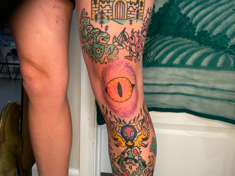 A photo of the front of my client’s leg shoing our in progress Lord of the Rings sleeve. The upper thing is eight months healed. The shin is around two years healed. The knee is fresh. In the center of the knee is the eye of Sauron. Around the knee is the script that appears around the one ring. 