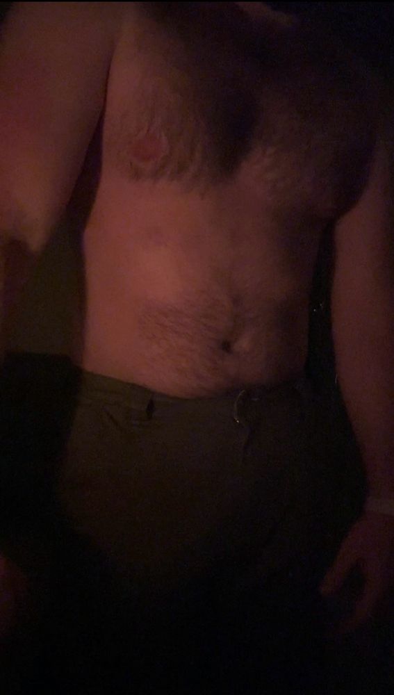 A shirtless, muscular, hairy person stands in a dark room lit by candlelight. We see them from the neck, down.