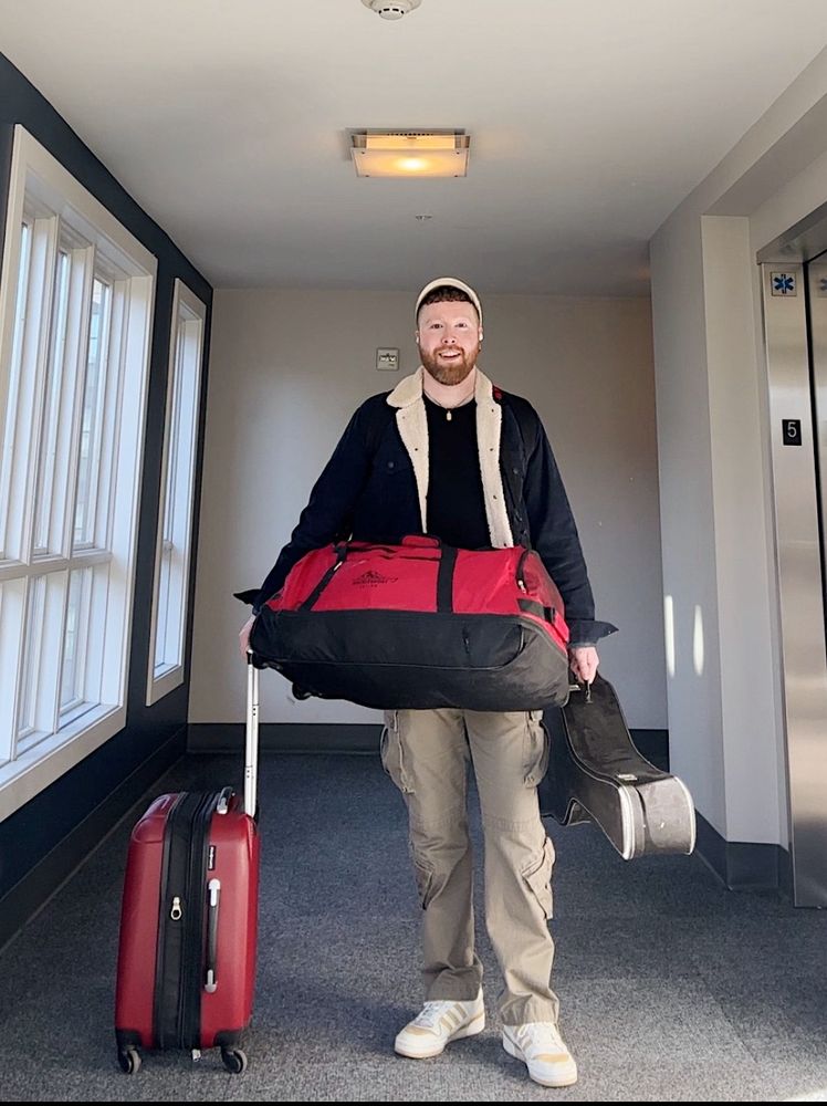 A bearded person stands in an elevator lobby with a red rolling suitcase, a large red duffel, and a guitar case. They are smiling.