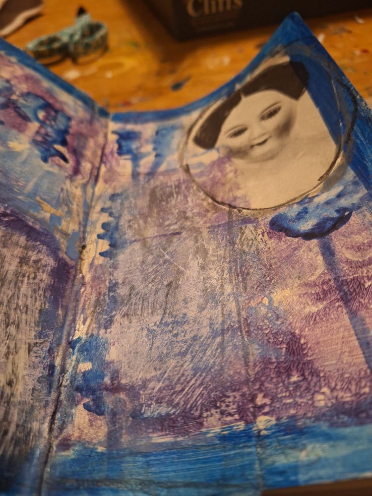Right side of a 2-page spread in a Dylusions art journal. The page is layered with book scraps, gesso, and smeared and drippy blue, purple, and silver paint. In the lower left corner, there is a scratchy white space that has a tall, shadowy mannequin body beneath it. In the upper right corner, there is a black + white picture of a black-haired porcelain doll face with dark blush and dark eyes. The page is bordered in dark blue.