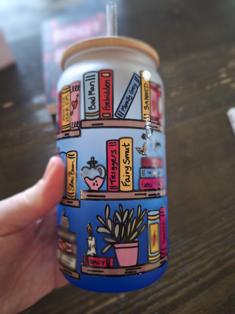 I'm holding a glass tumbler with a wooden top and plastic straw. It has hand drawings of bookshelves with books, plants, candles, and potion bottles. The bookshelves titles depicting tropes in smut & romance novels: Bad Man, Forbidden, Morally Grey, Banned, Fairy Smut, etc.