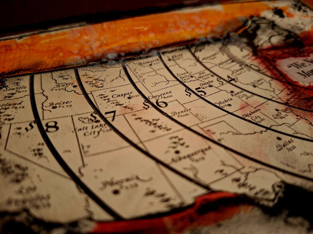 Close up of my art journal page. Layered on top of orange and red acrylic paint is a vintage map of the United States. The map has zones marked with prices listed each zone, getting less expensive from West to East. The map is outlined in black pastels.
