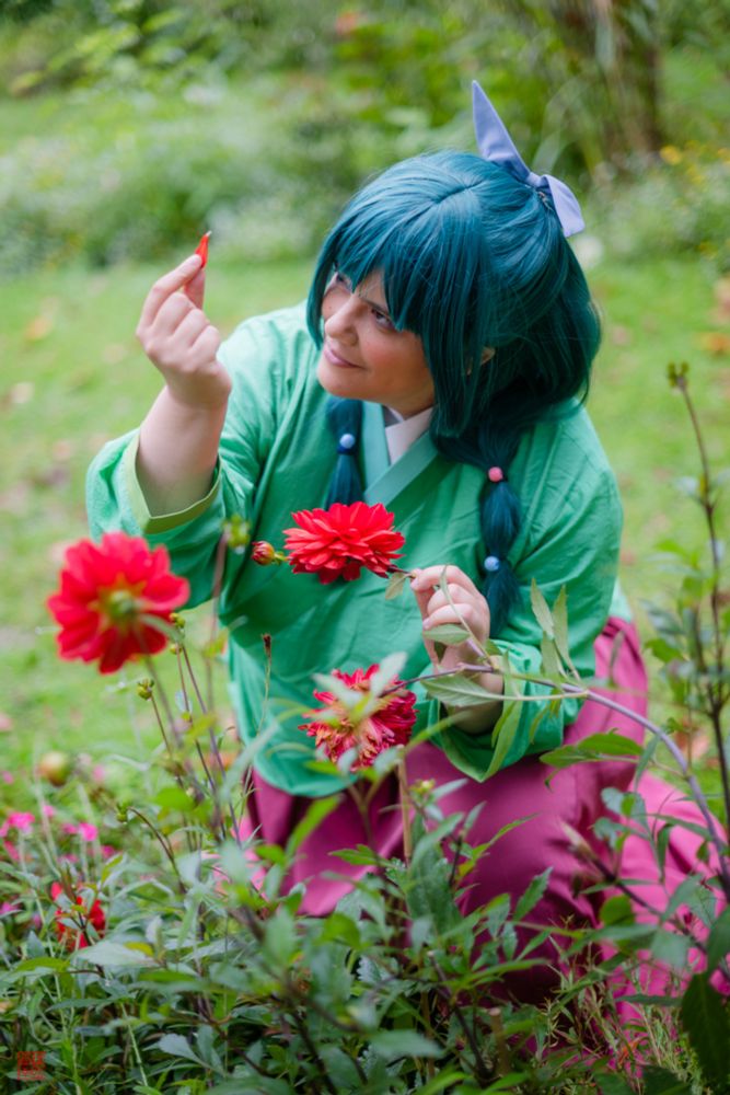 Illiane cosplaying as Mao Mao from the Apothecary's Diaries (in her classical green/red hanfu). She's in a middle of a garden, holding a red flower in one hand and a petal of the same flower in the other hand. She's looking at the petal with curiosity and excitement 