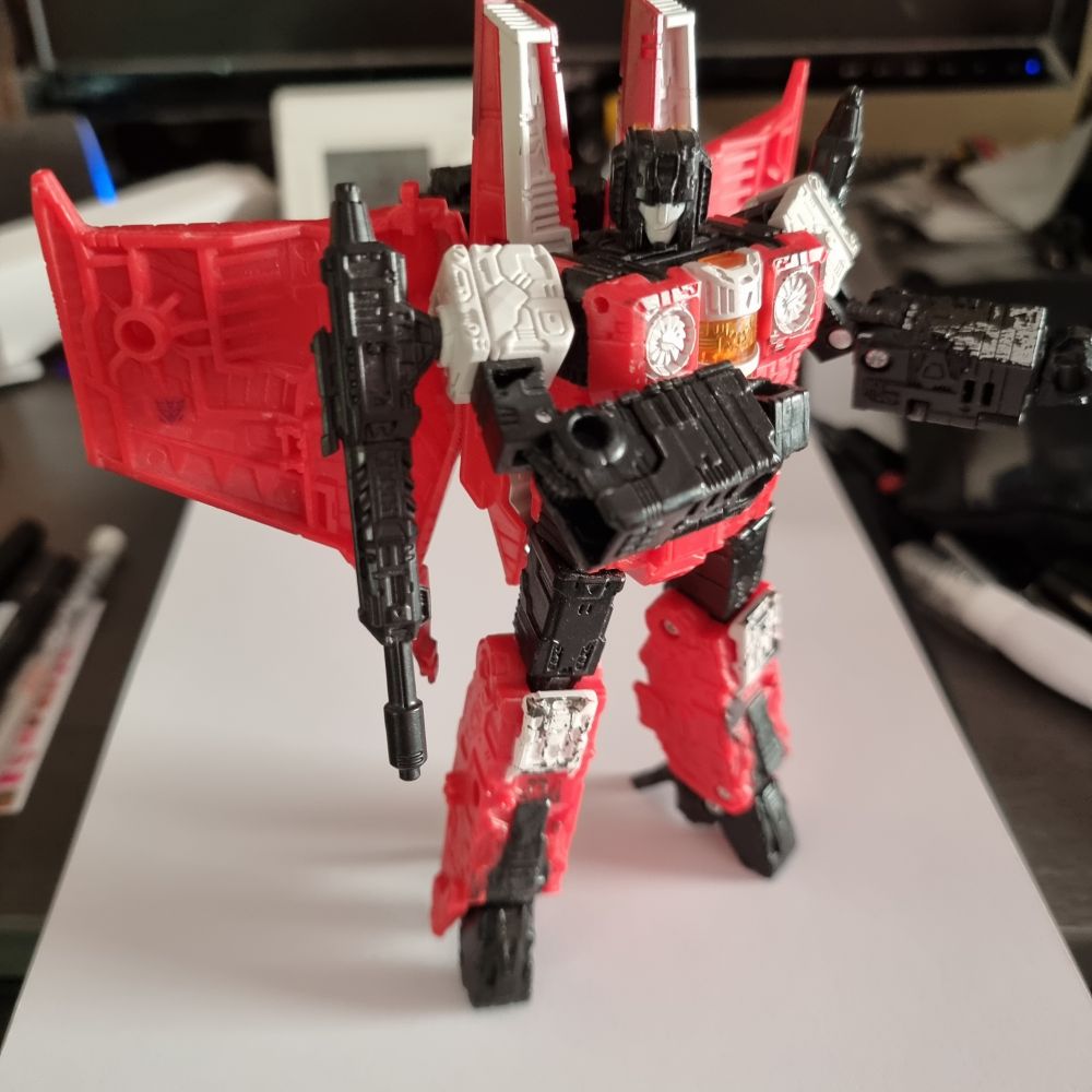 Seige Red Wing in robot mode standing on an open sketchbook on a cluttered desk. 