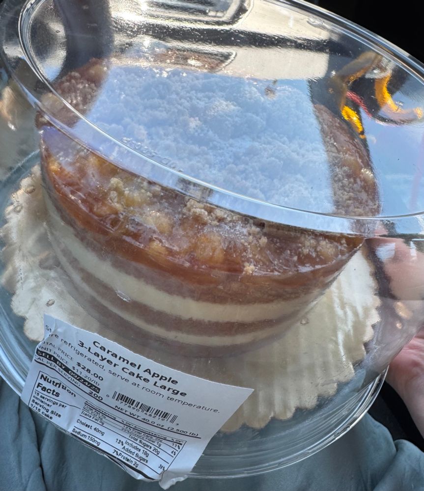 a photo someone took in a car of a cake from wegman’s. The label says “caramel apple 3-layer cake large”. It has apple caramel cinnamon goo on top