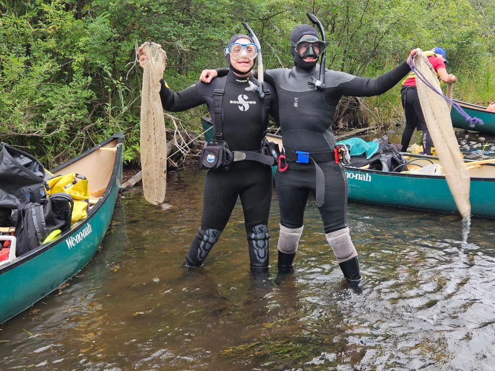 Two women in SCUBA gear standing in a river next to a canoe