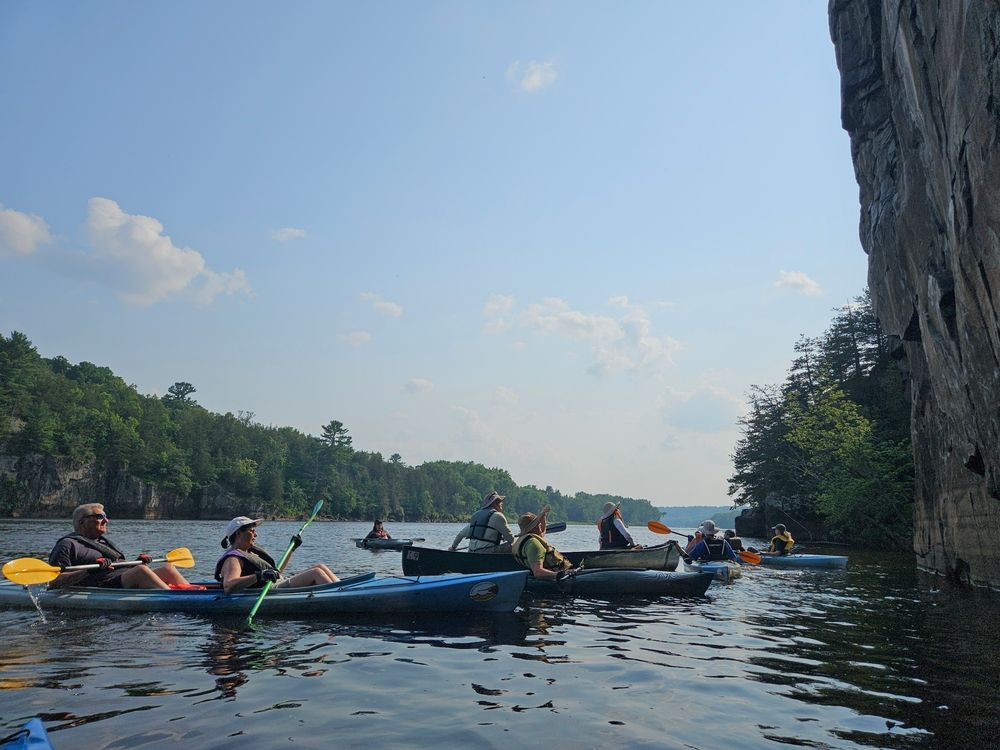 People in kayaks and a canoe on a river in front of a tall basalt cliff