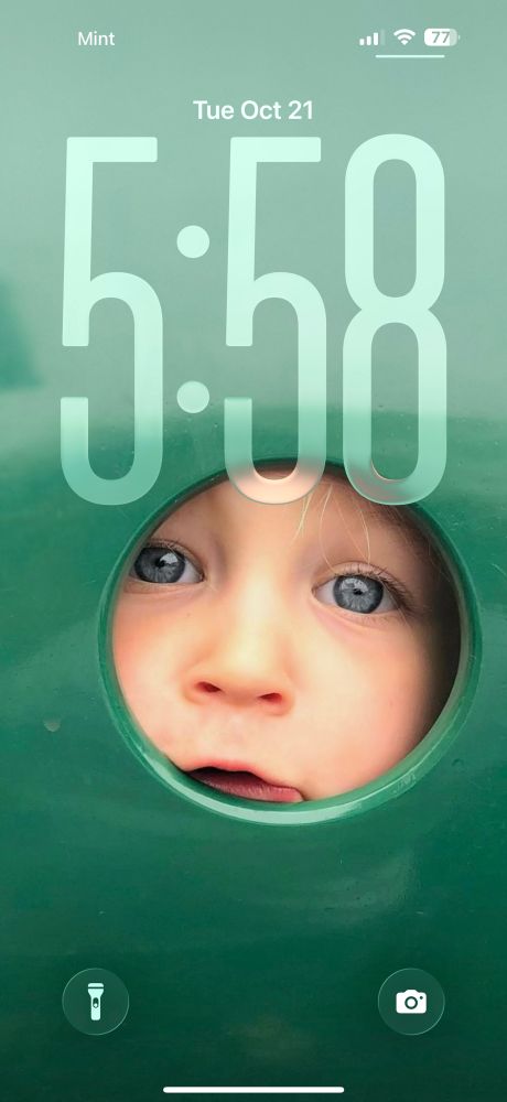A little girl peeks through a hole in a green piece of playground equipment 