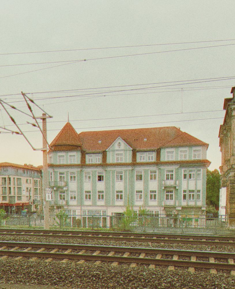 Ein Haus im Hintergrund und im Vordergrund führen Schienen entlang. Der Himmel ist Wolkenverhangenen. Das Bild hat einen Filter, der der Analogphotographie nachahmt. 