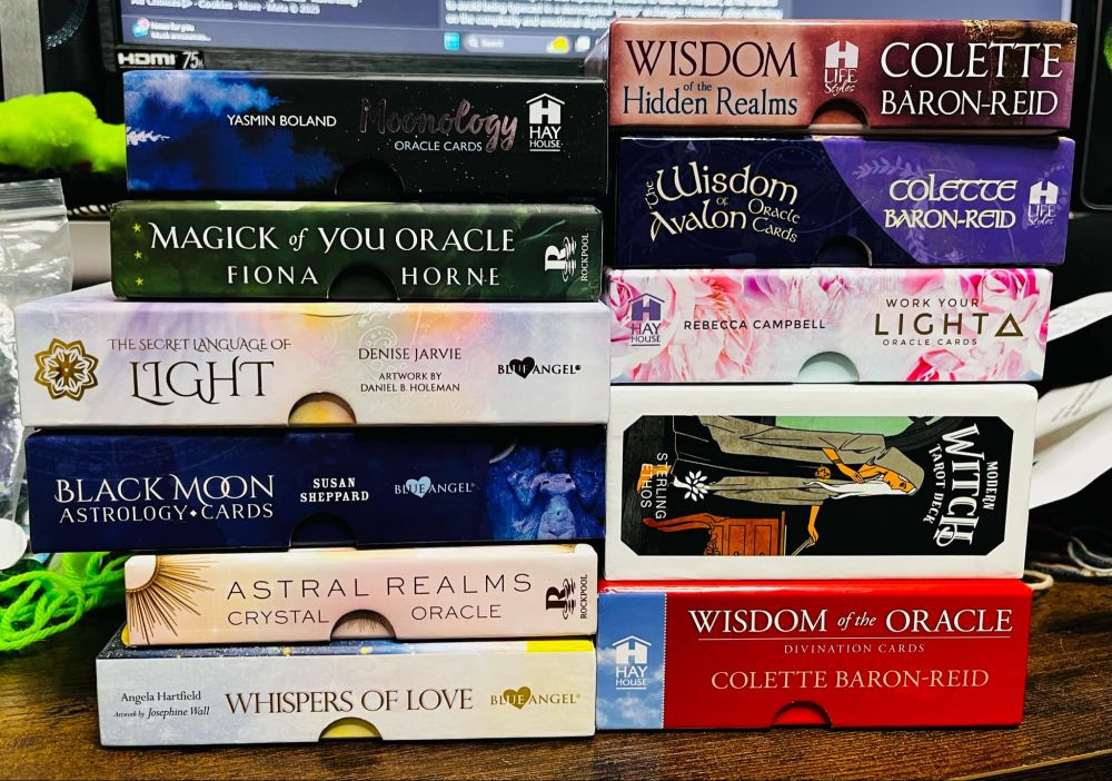A collection of 11 oracle and tarot decks stacked in two columns on a wooden surface. The left stack includes Moonology Oracle Cards by Yasmin Boland, Magick of You Oracle by Fiona Horne, The Secret Language of Light by Denise Jarvie, Black Moon Astrology Cards by Susan Sheppard, Astral Realms Crystal Oracle, and Whispers of Love by Angela Hartfield. The right stack features Wisdom of the Hidden Realms, The Wisdom of Avalon, and Wisdom of the Oracle by Colette Baron-Reid, Work Your Light Oracle Cards by Rebecca Campbell, and Modern Witch Tarot Deck by Sterling Ethos. A computer monitor is visible in the background, along with various objects scattered around, creating a cozy, mystical ambiance.