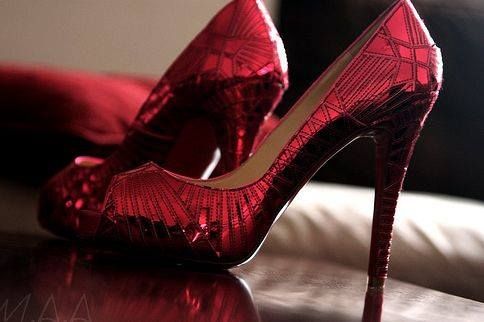 A pair of shiny, metallic red high-heeled shoes with a pointed toe and stiletto heel. The surface features a geometric, almost art deco pattern that reflects light, giving them a glossy and eye-catching appearance. They are placed on a smooth surface with a blurred background of cushions.